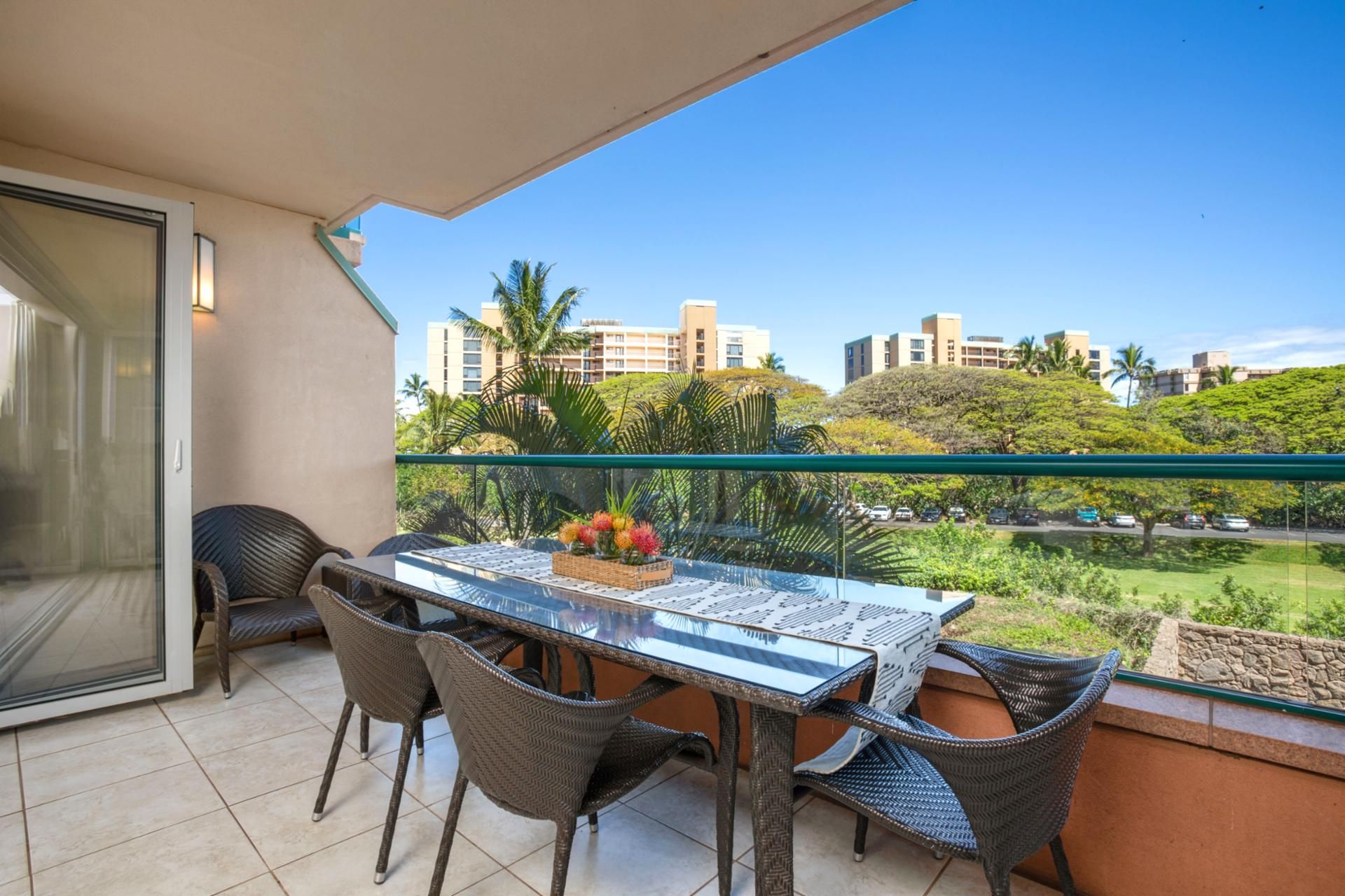 A balcony with a glass table and wicker chairs, overlooking a residential area with palm trees and buildings.
