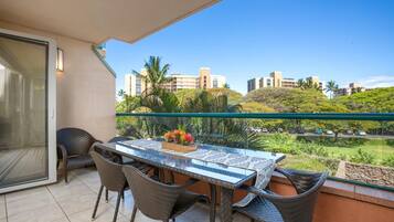A balcony with a glass table and wicker chairs, overlooking a residential area with palm trees and buildings.