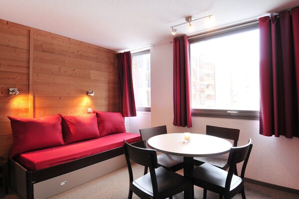 A compact room with a wooden wall, a red sofa, a round dining table with four chairs, and large windows with red curtains.