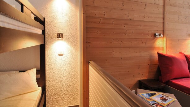 Habitación de hotel con literas, pared de madera, una mesita con revistas y una almohada roja en un sofá.