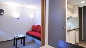 A compact room with a bed, a small table, a bench, a wall-mounted TV, and a kitchenette area. - travelski home select - Résidence Les Glaciers - Studio 3/4 personnes 318 (La Plagne)