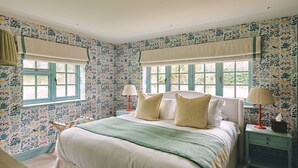 A bedroom with floral wallpaper, a bed with white bedding, two bedside tables with lamps, and a window with a curtain. - Bridlepath Cottage - Luxury Cottages (Hungerford)