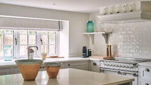 A kitchen with a large window, white cabinets, a stove, and a coffee maker. - Bridlepath Cottage - Luxury Cottages (Hungerford)