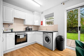 A modern kitchen with white cabinets, a built-in oven, a washing machine, and a large window with a view of a green garden.
