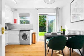 A modern kitchen with a washing machine, a dining area with a table and chairs, and a view of a garden through a sliding glass door.