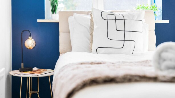 Ein Schlafzimmer mit blauer Akzentwand, einem Nachttisch mit Lampe, einem Fenster mit Ausblick und einem Bett mit weißer Bettwäsche und einem geometrischen Kissen.
