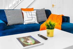 A blue sofa with patterned cushions, a white table with a remote control and a plant, and a door in the background.