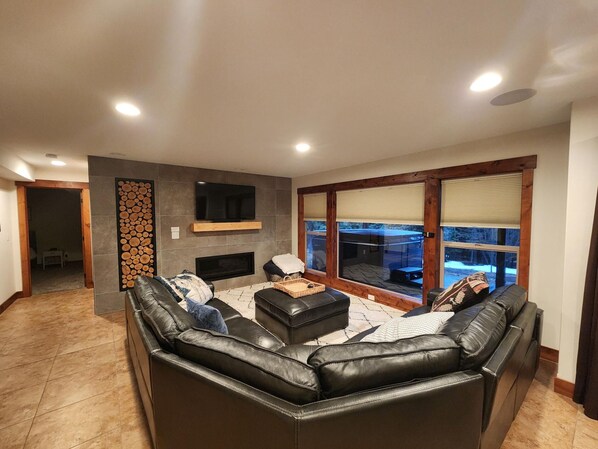 A modern living room with a fireplace, leather sofas, a TV, and large windows. - The Haskill Highland Retreat (Whitefish)