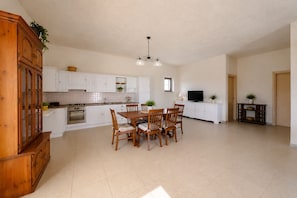 A spacious kitchen with white cabinets, a dining table with chairs, a wooden cabinet, a television on a white cabinet, and a hanging light fixture.