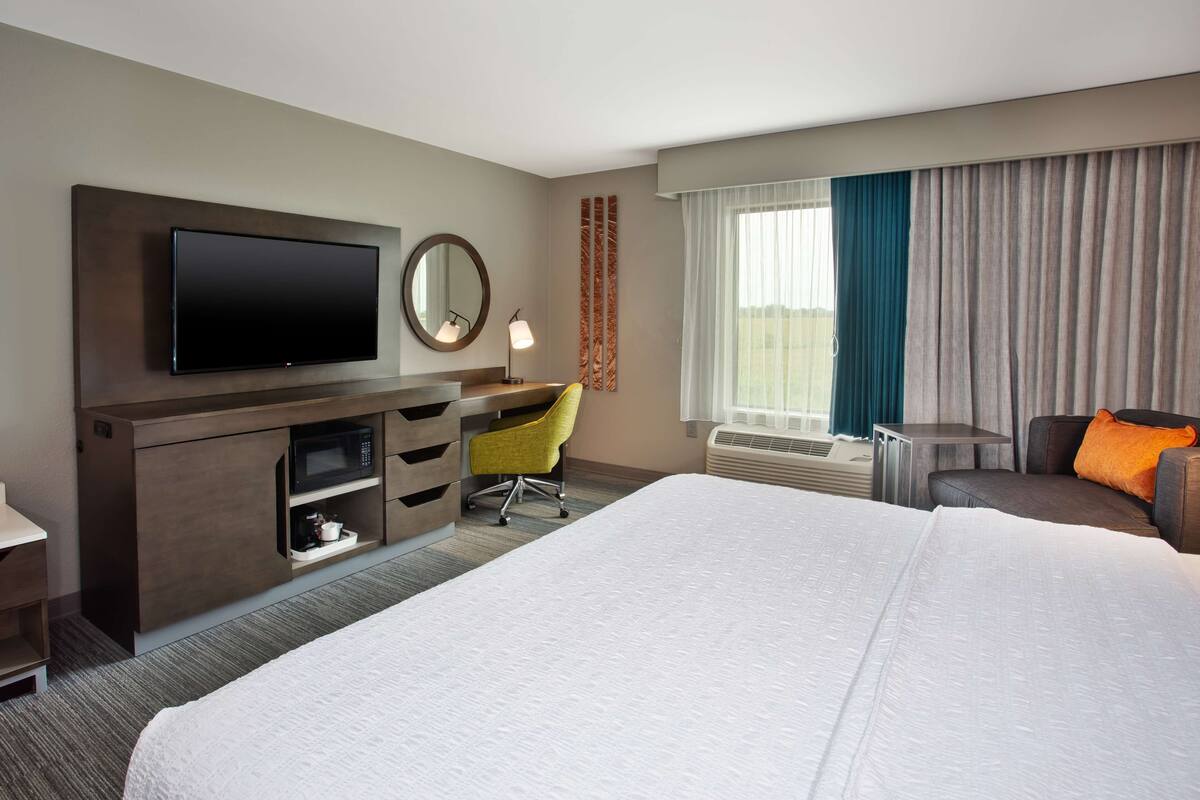 A hotel room with a large bed, a desk with a chair, a flat-screen TV, a mirror, and a window with curtains.