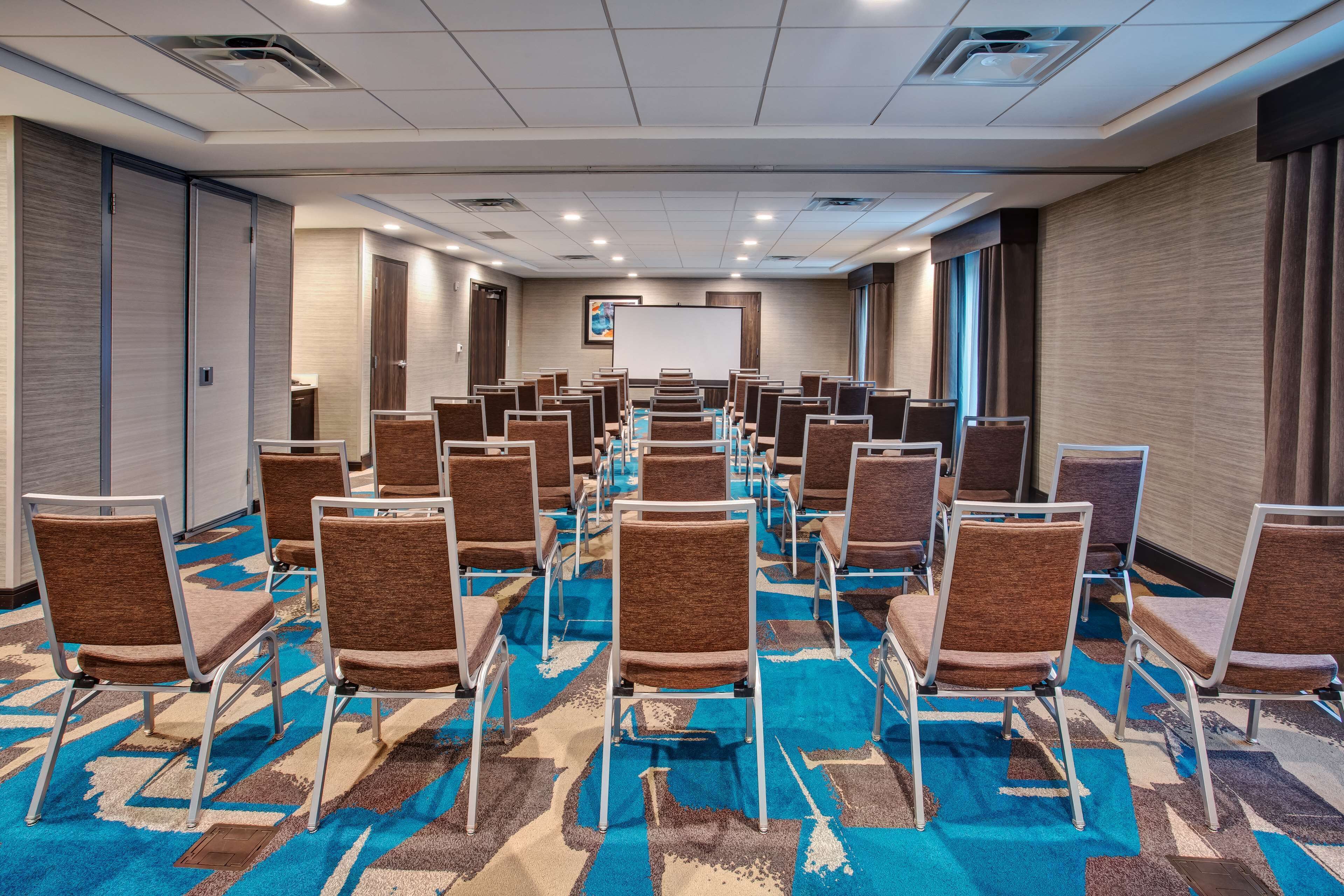 Un salón de conferencias con filas de sillas, una pantalla de proyector y piso alfombrado.