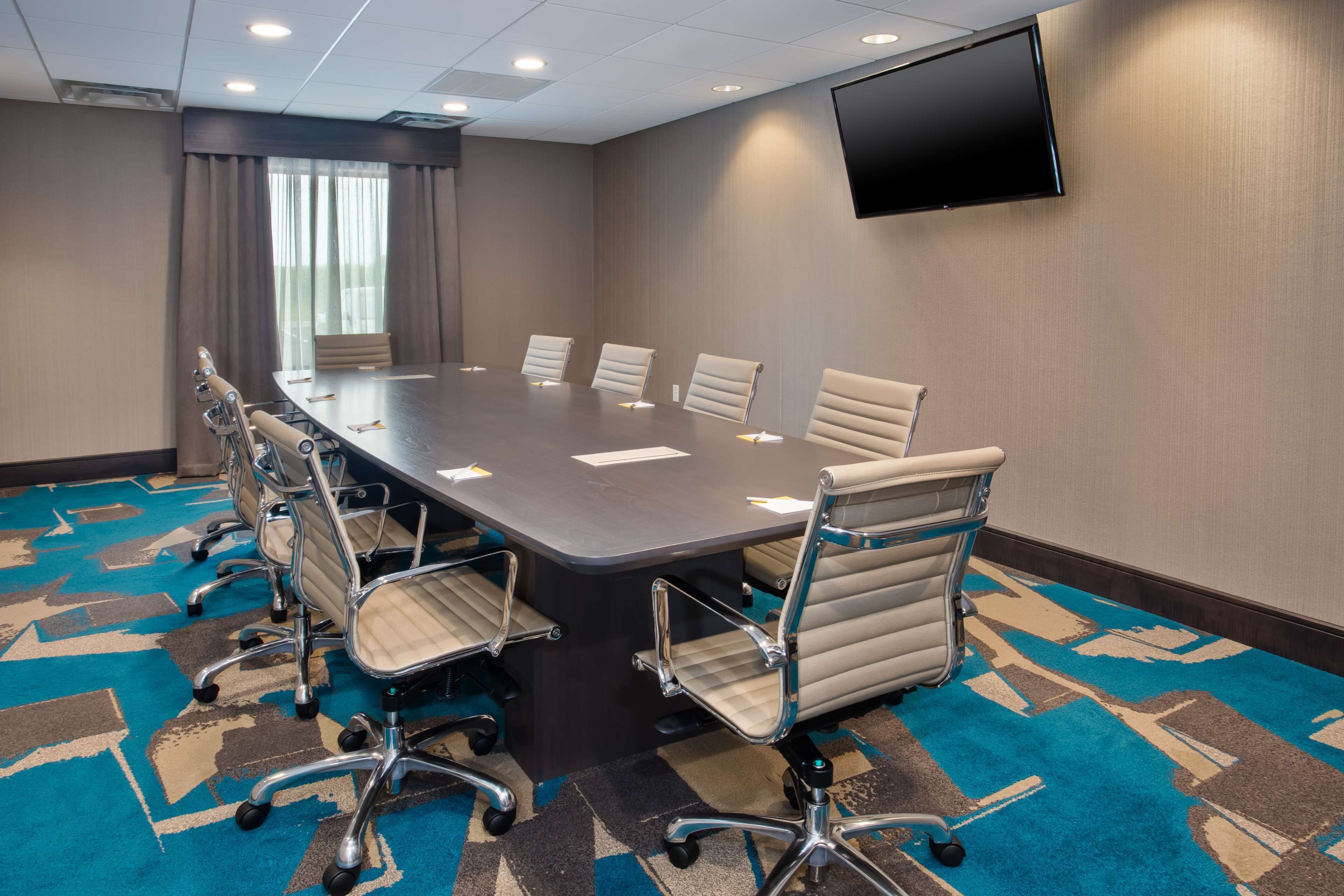 A conference room with a long table, six chairs, a flat-screen TV, and a patterned carpet.