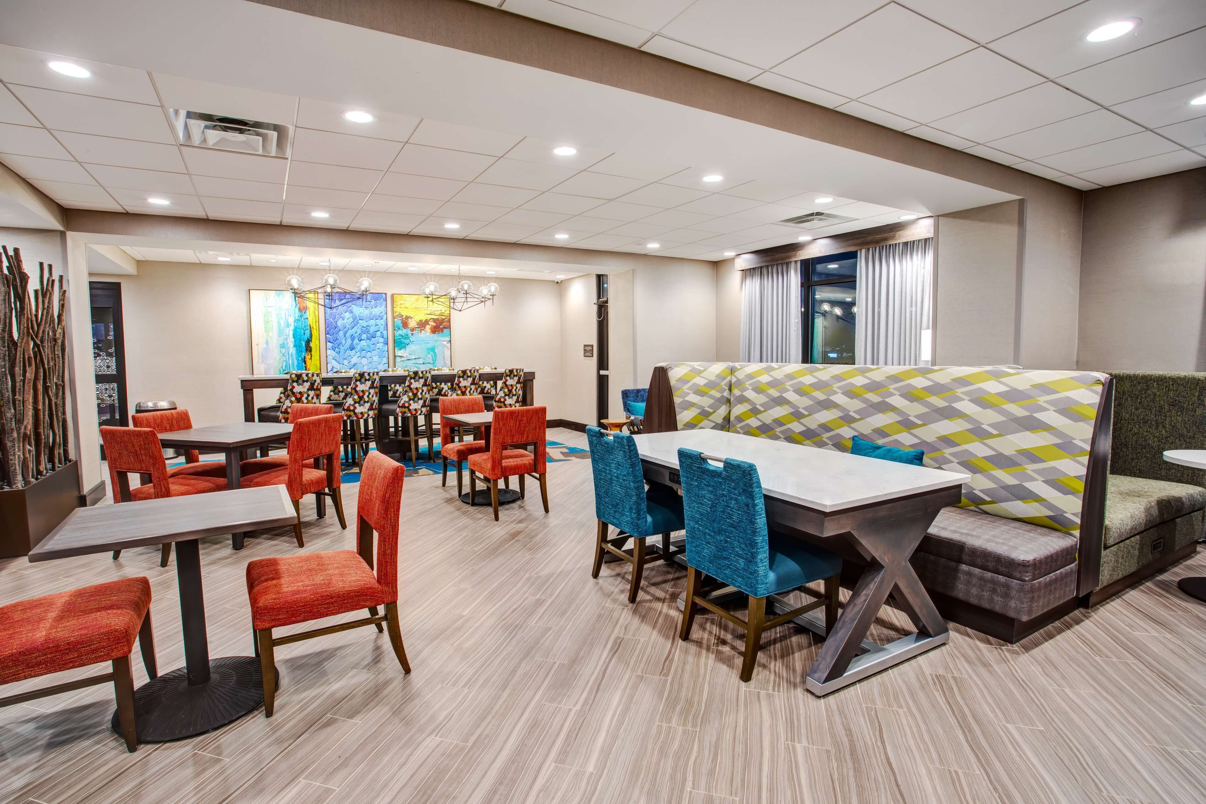 A hotel dining area with multiple tables and chairs, a central seating area with a patterned cushion, and artwork on the walls.