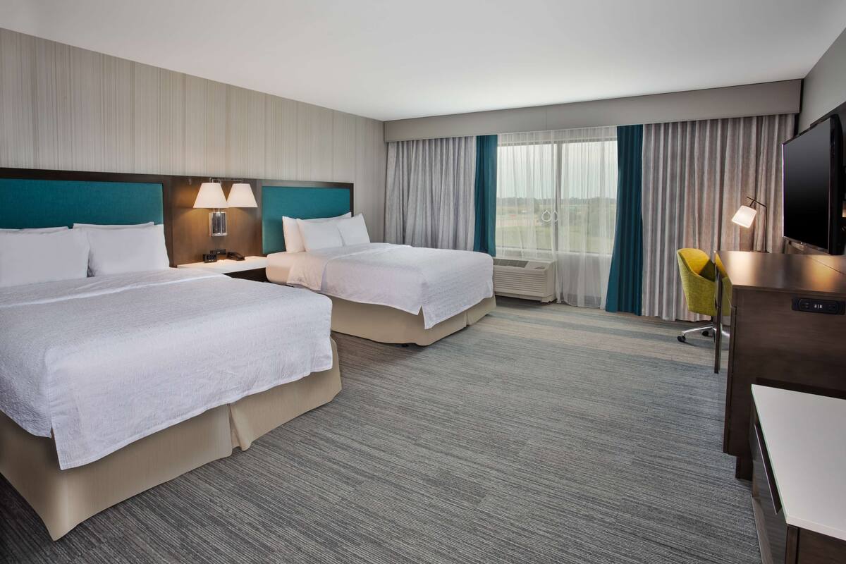 A hotel room with two beds, a desk, a TV, and a window with curtains.