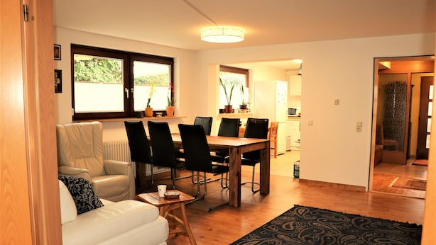 Un salon avec un canapé blanc, des fauteuils noirs, une table à manger en bois et un tapis à motifs.