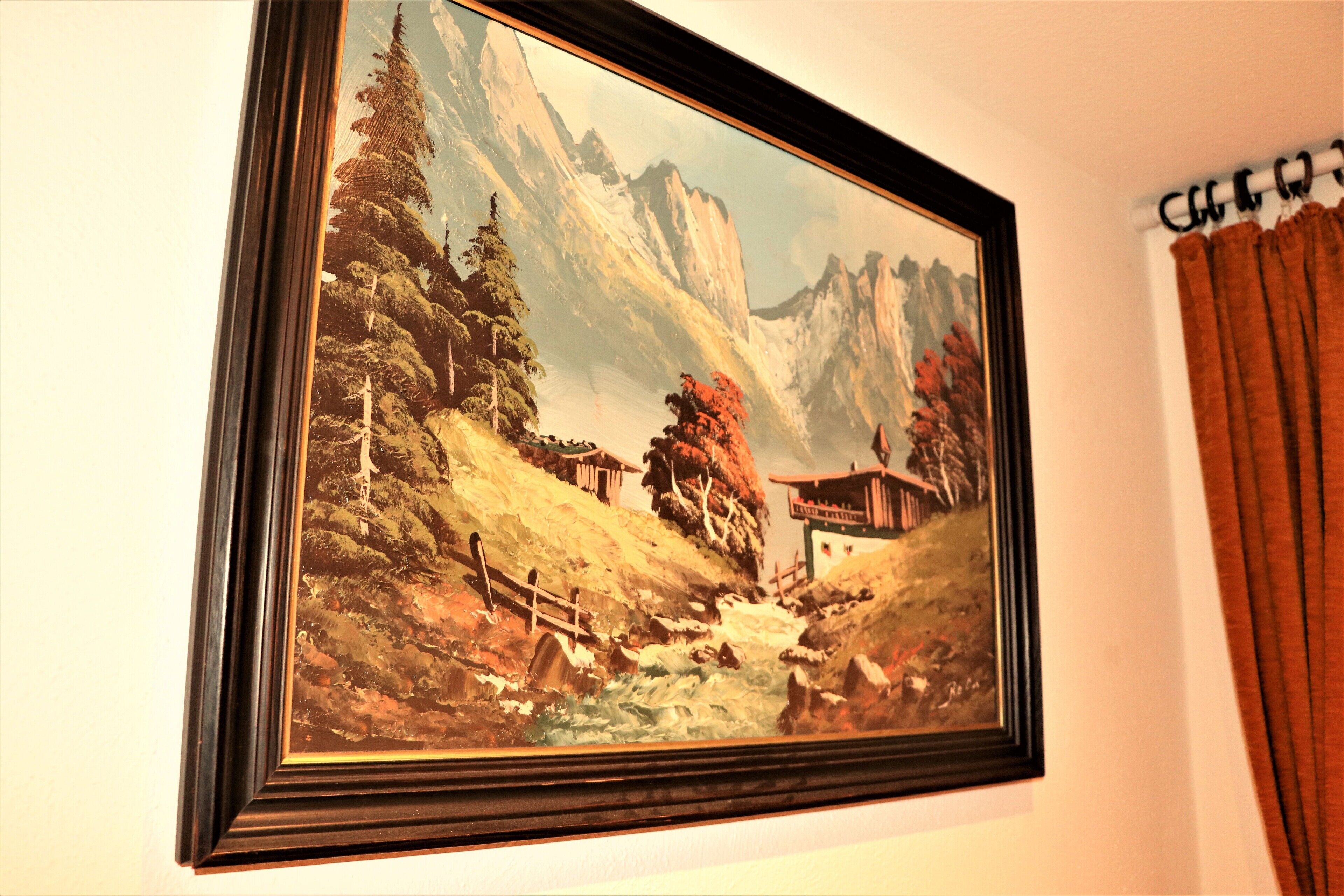 A framed painting of a mountainous landscape with a wooden cabin and a bridge.
