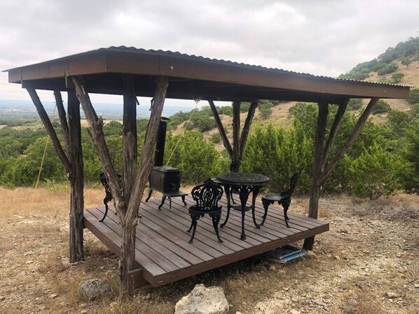 Une terrasse en bois surmontée d’un toit en métal, entourée d’arbres et de collines.