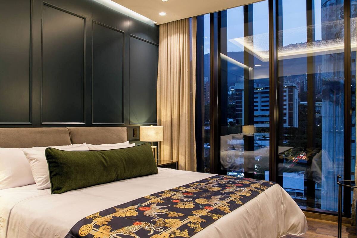 A hotel room with a large bed, a city view through floor-to-ceiling windows, and dark paneling on the walls.