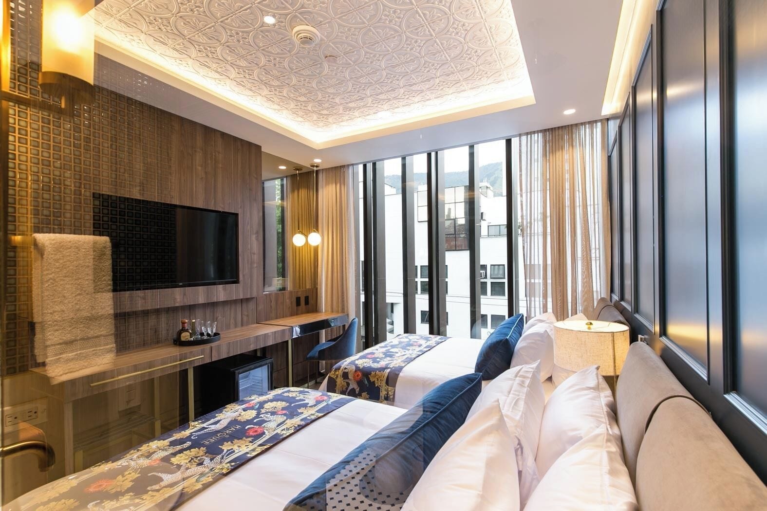 A modern hotel room with a large bed, a desk, a TV, and a view of the city through large windows.
