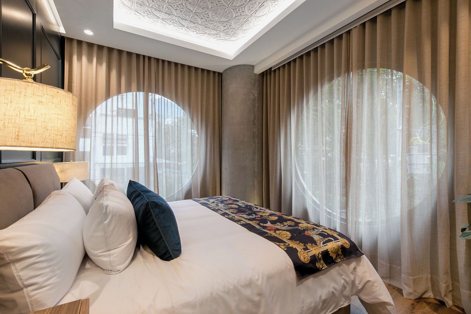 A modern bedroom with a bed, a lamp, and large windows with curtains.