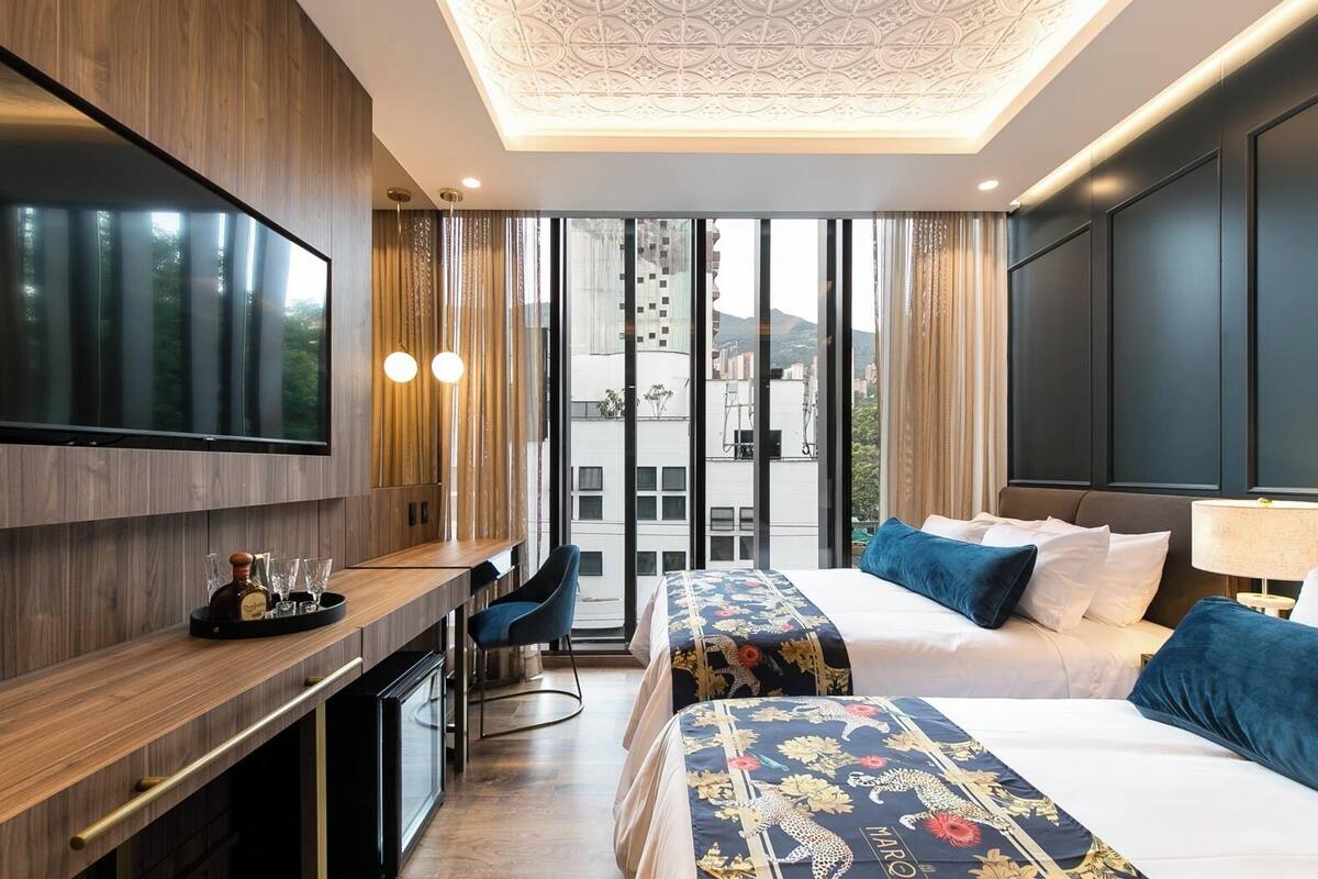 A hotel room with a large TV, a desk with a bottle and glasses, two beds with patterned bedspreads, and a view of buildings through large windows.