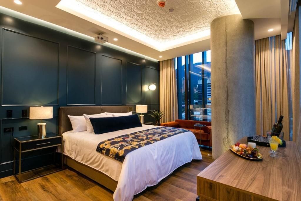 A modern hotel room with a large bed, a wooden side table with a drink and fruit, and a view of the city through large windows.