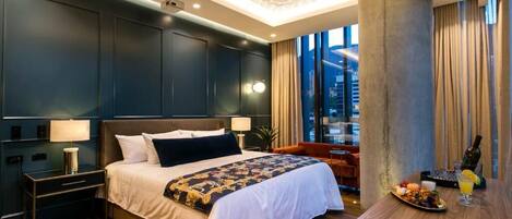 A modern hotel room with a large bed, a wooden side table with a drink and fruit, and a view of the city through large windows.