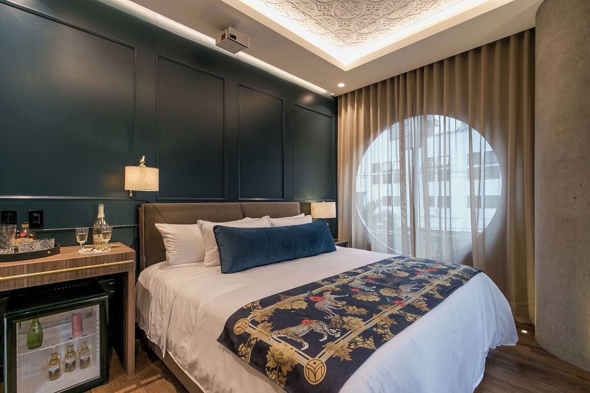 A hotel room with a large bed, a nightstand with a lamp, a minibar, and a large window with curtains.