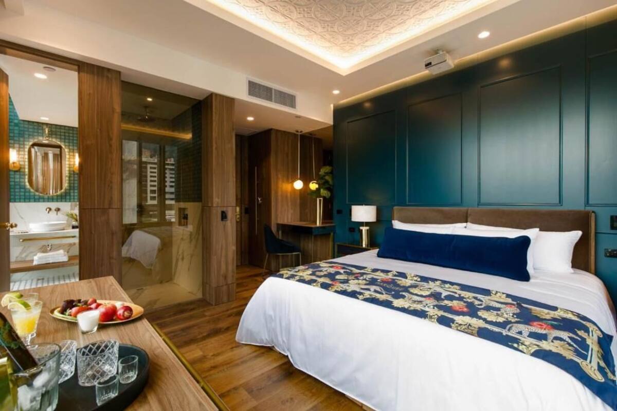 A modern hotel room with a large bed, a wooden desk with a fruit plate, a bathroom with a mirror and sink, and a teal accent wall.
