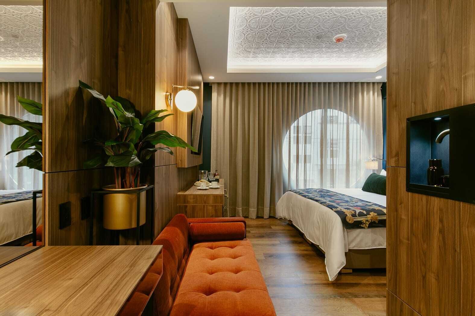A modern hotel room with a wooden floor, a large plant, a round table, a sofa, a bed with a white bedspread, a TV mounted on the wall, and a ceiling with a decorative pattern.
