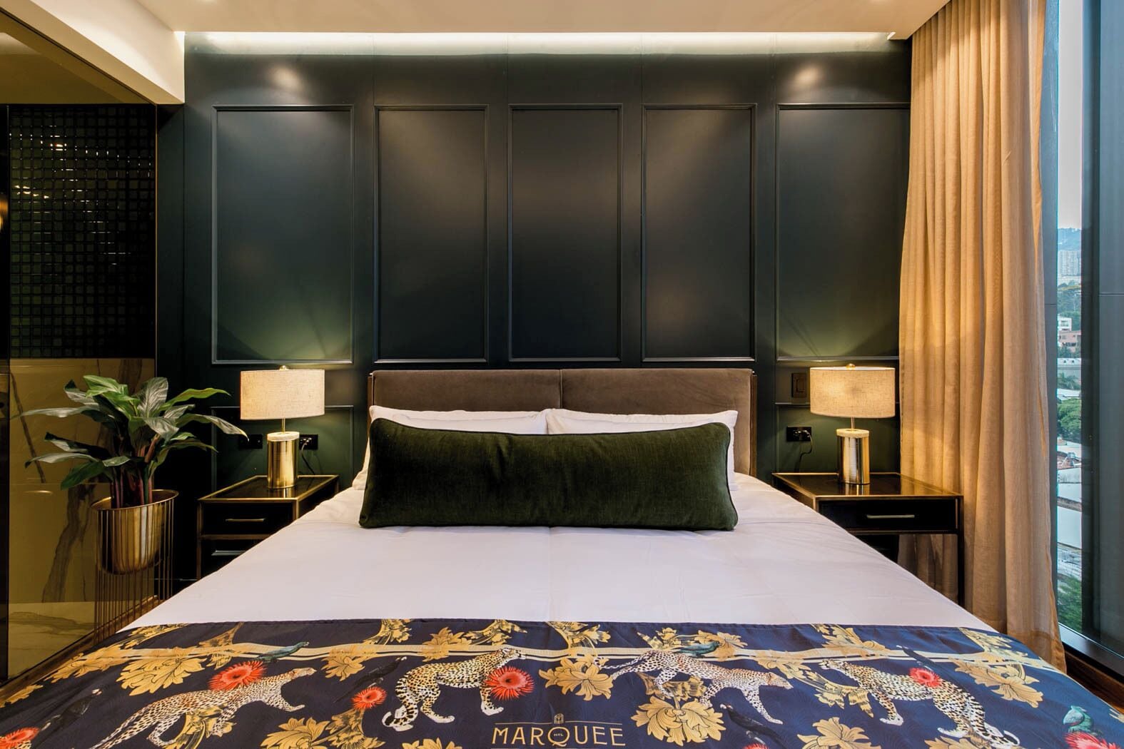 A modern hotel room with a large bed, dark paneled walls, and a view of the city through large windows.