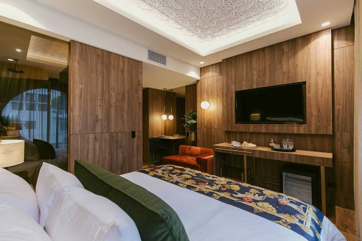 A modern hotel room with a large bed, a TV mounted on the wall, a small desk with a chair, and a seating area with a sofa and armchair.