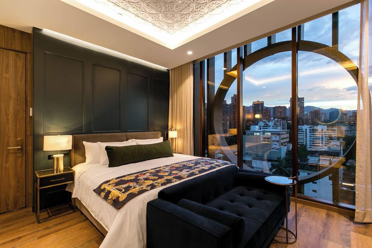 A modern hotel room with a large bed, a nightstand, a reading chair, and a city view through floor-to-ceiling windows.