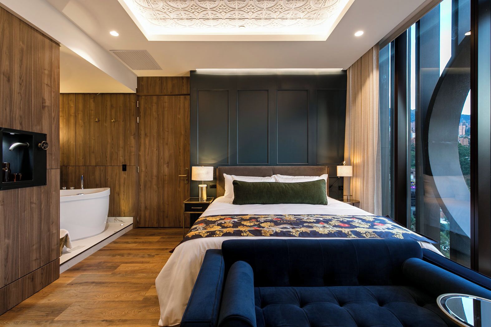 A modern hotel room with a large bed, a built-in bathtub, and a view of the city through large windows.