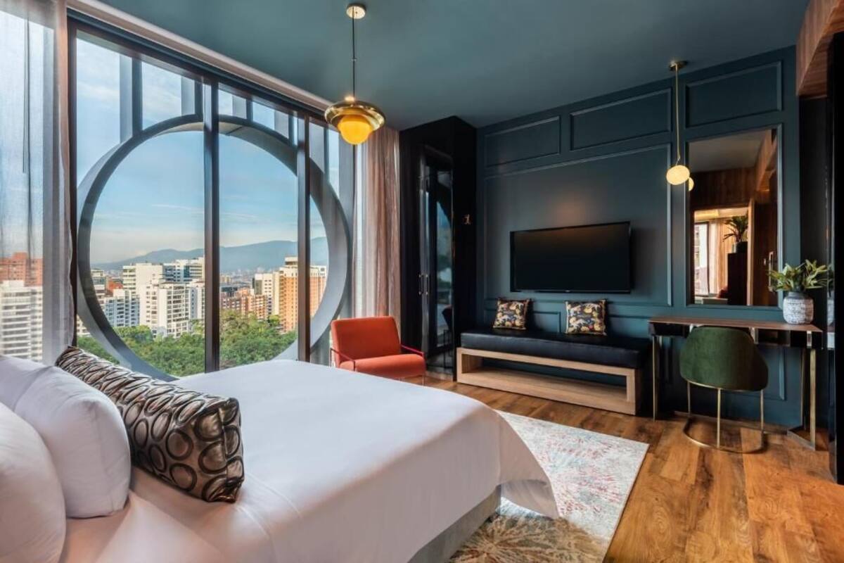 A modern hotel room with a large bed, a desk, a TV, and a view of the city.