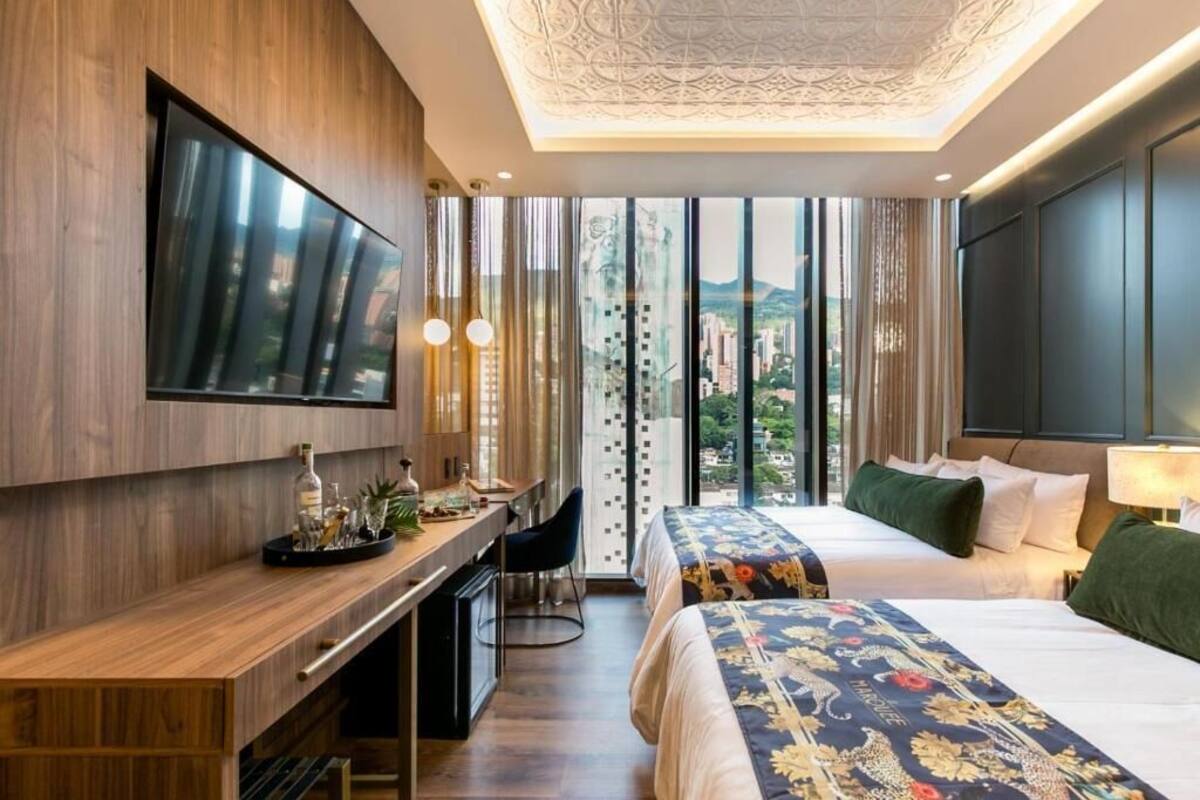 A hotel room with a large bed, a desk with a chair, a TV mounted on the wall, and a view of the city through large windows.