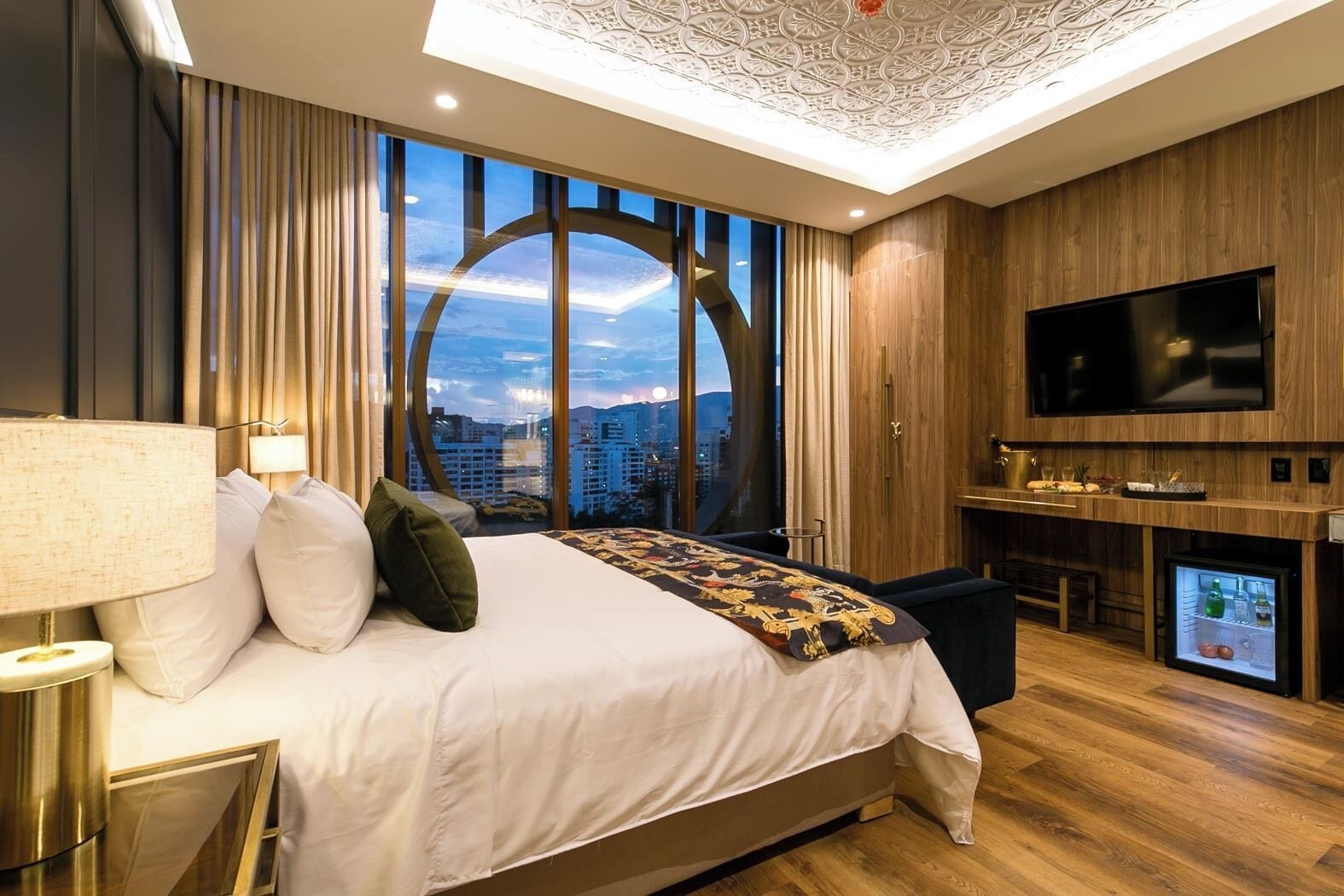 Una habitación de hotel moderna con una cama grande, televisión, refrigerador minibar y vista a la ciudad desde la ventana.