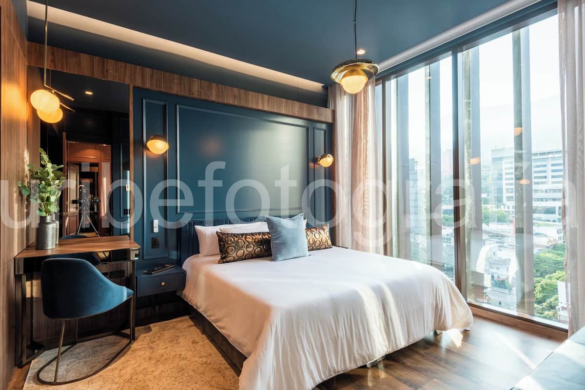 A modern bedroom with a large bed, a desk, and a chair. There is a view of the city through large windows.