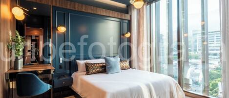 Un dormitorio moderno con una cama grande, un escritorio y una silla. Se ve la ciudad a través de ventanas amplias.