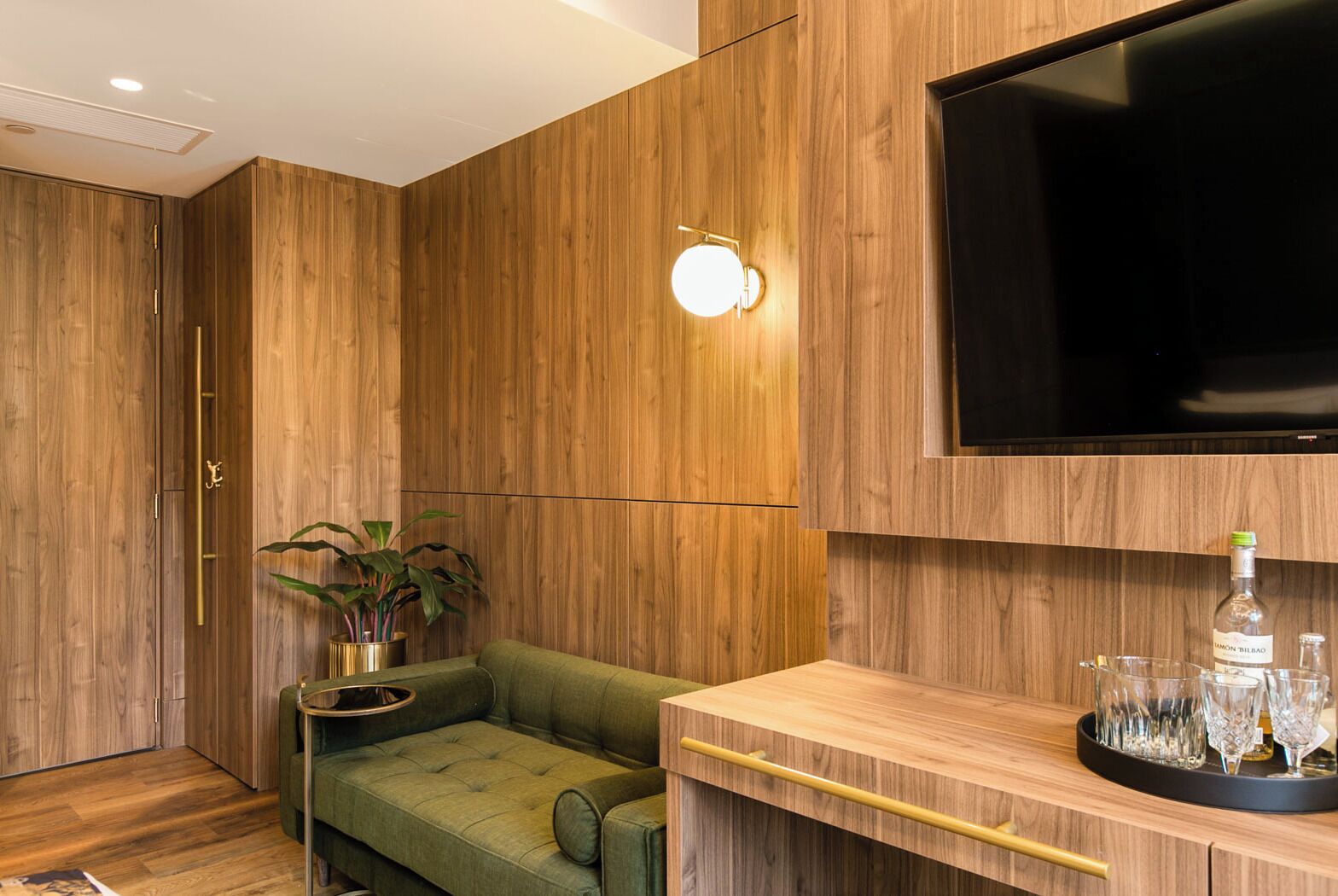 A modern living room with a green sofa, a wooden TV stand, a flat-screen TV, and a small table with a plant and a tray of glasses.