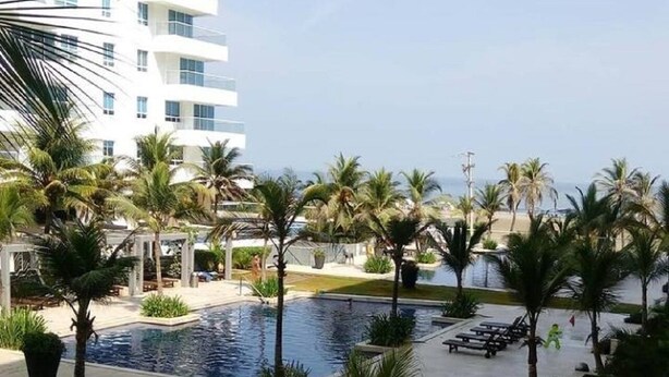 A pool area with palm trees, lounge chairs, and a modern building in the background. - 1ME2 Apartamento en Morros Frente al Mar (Cartagena)