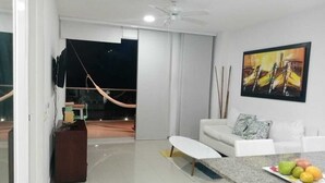 A modern living room with a white sofa, a coffee table, and a painting on the wall. There is a balcony with a hammock and a view of the outside. - 1ME2 Apartamento en Morros Frente al Mar (Cartagena)