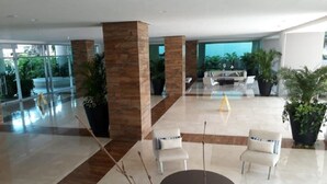 A spacious lobby with a modern design, featuring a central seating area with white chairs and a round table, surrounded by large potted plants and a glass wall offering a view of the outdoors. - 1ME2 Apartamento en Morros Frente al Mar (Cartagena)
