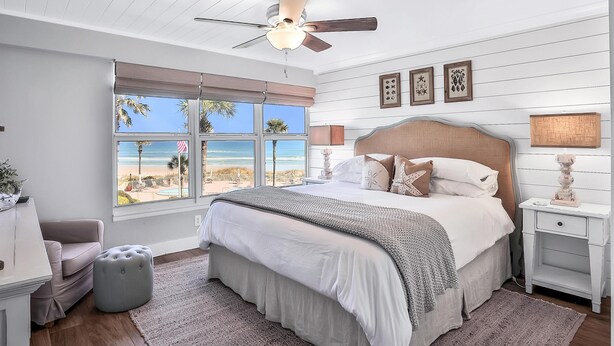 A bedroom with a large bed, a ceiling fan, a nightstand, a lamp, and a view of the beach. - Picturesque, High End Vaca Condo, Atlantic Views from all corners. Sunset Deck  (St. Augustine)