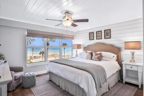 Et soveværelse med en stor seng, ventilator i loftet, vindue med udsigt til stranden og et natbord med lampe.