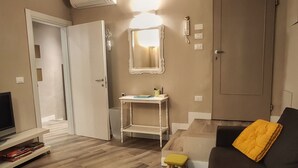 Een woonkamer met een houten vloer, een grijze bank met een geel kussen, een klein krukje, een witte tafel met een spiegel, en een deur die naar een andere kamer leidt.