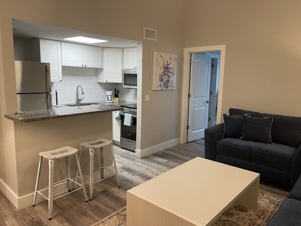 A modern kitchen with white cabinets, a stainless steel refrigerator, and a microwave. There is a dining area with a white table and two bar stools. A dark-colored sofa is in the living room, and a door leads to another room. - Siesta Key Village-Suite #4: A block to the Beach! (Sarasota)