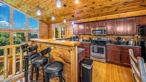 A wooden cabin kitchen with a bar area, bar stools, and a large window overlooking greenery.