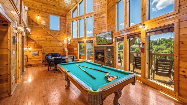 A wooden interior with a pool table, seating area, and large windows offering views of the outdoors.