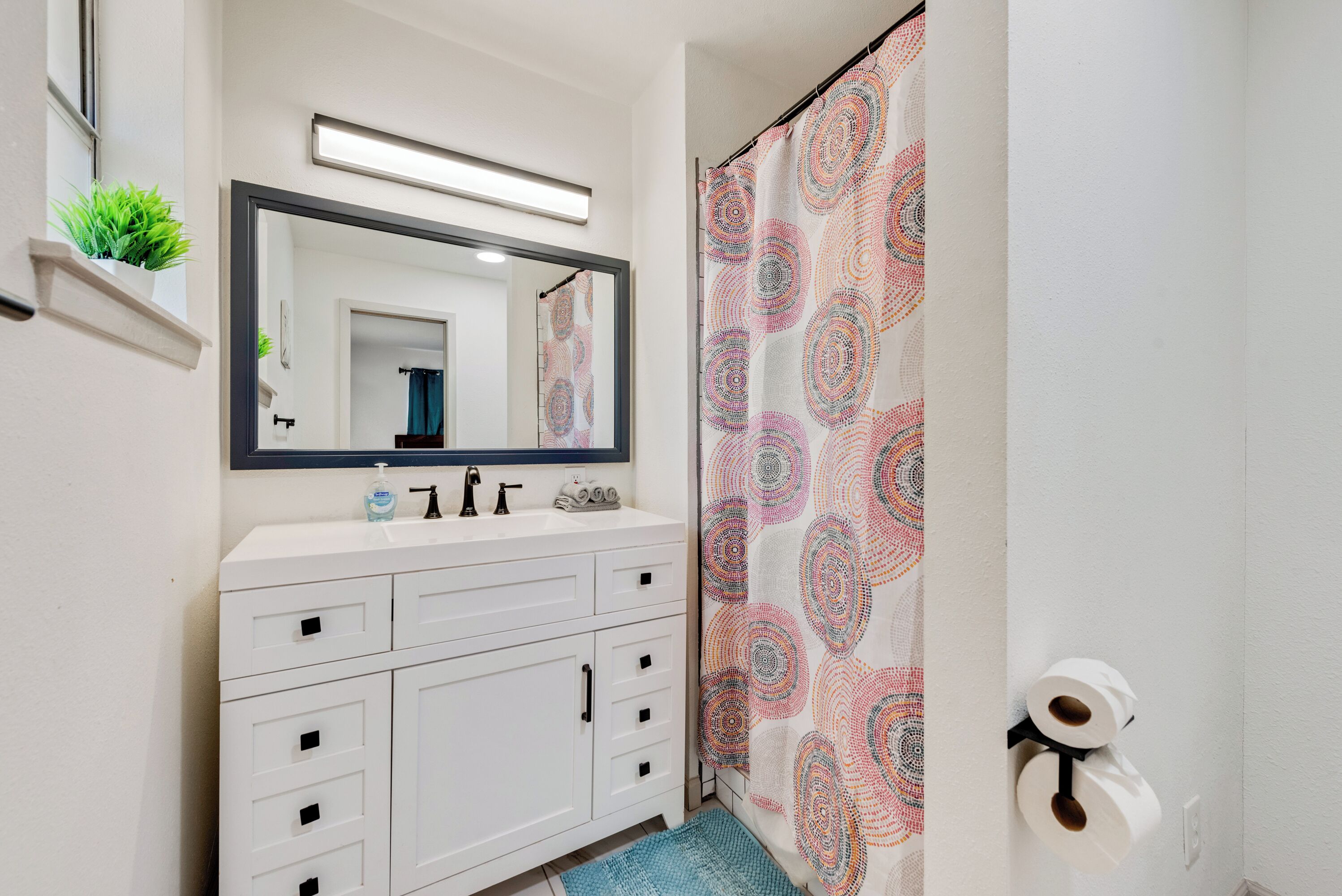 Une salle de bain avec un meuble-lavabo blanc, un miroir, une étagère avec une plante et un rideau de douche orné d’un motif circulaire.